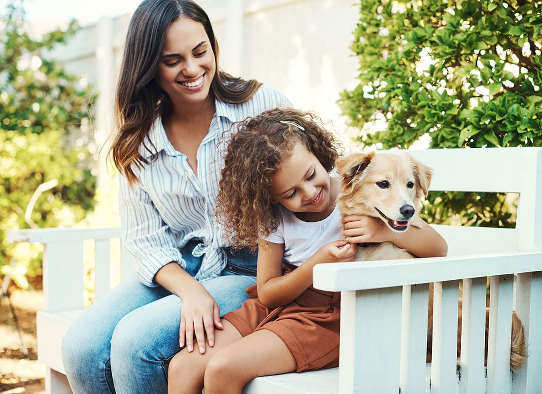 Personal Insurance - Smiling Mom and Daughter Sitting on a White Bench with Their Dog in the Park on a Sunny Day
